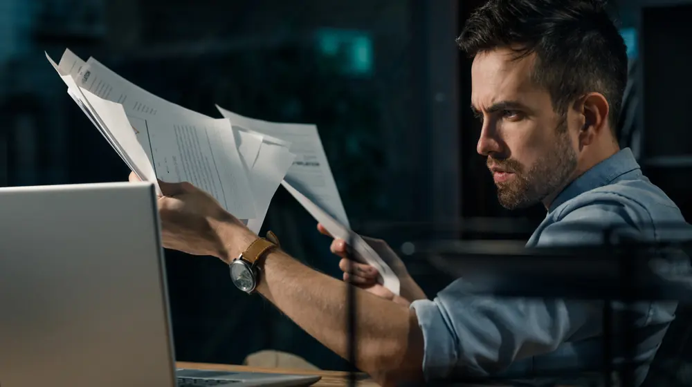 busy business owner holding papers in his hand while looking at a laptop screen.